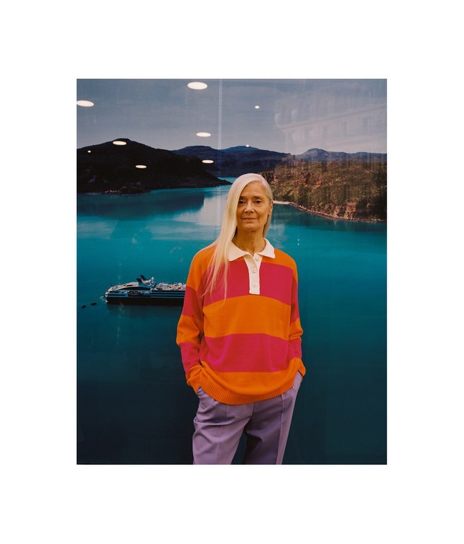 Older woman with long gray hair wears a pink and orange striped shirt and light purple pants in front of a scenic lake photograph.