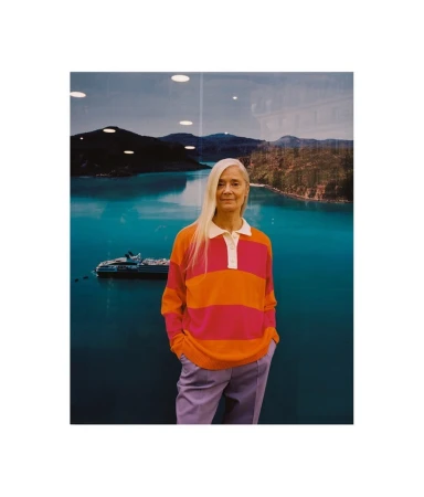 Older woman with long gray hair wears a pink and orange striped shirt and light purple pants in front of a scenic lake photograph.