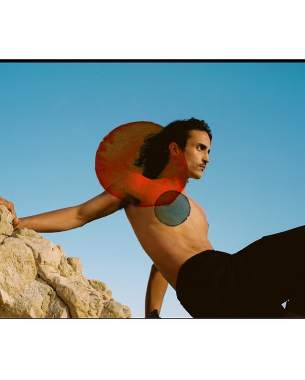 Man with dark hair leaning against rock under blue sky with overlaid red and blue circular watercolor marks
