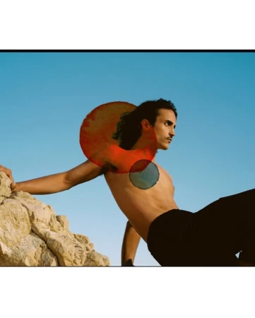 Man with dark hair leaning against rock under blue sky with overlaid red and blue circular watercolor marks
