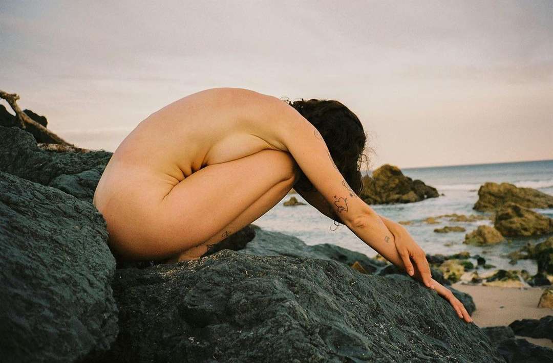 Nude woman crouches on rocks by ocean. Close up shot of brunette, dark hair, tattoos, sitting hunched at beach with rocks and water.