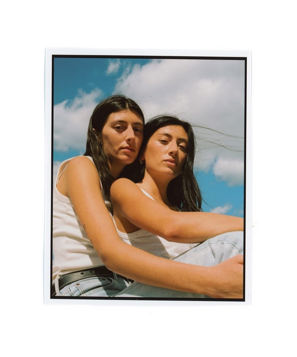 Two women pose closely together outdoors against a backdrop of a blue sky with clouds, both wearing casual tank tops and light jeans.