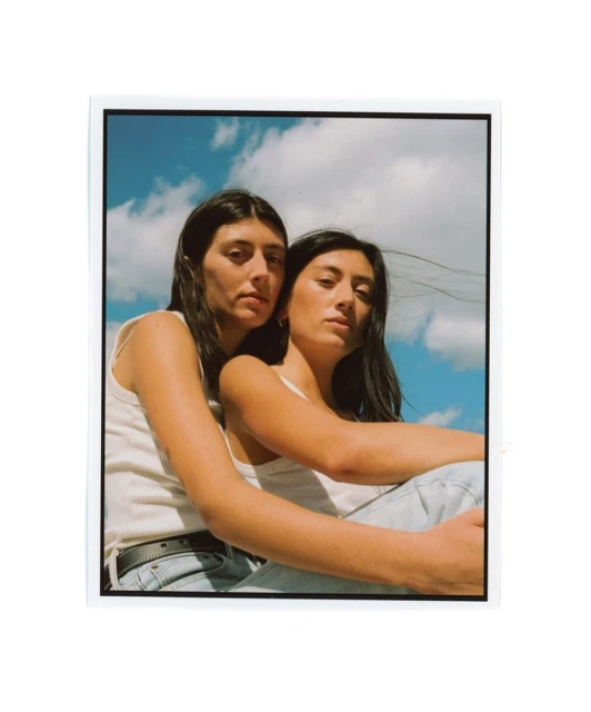Two women pose closely together outdoors against a backdrop of a blue sky with clouds, both wearing casual tank tops and light jeans.