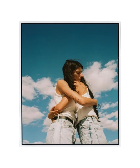 Two women embrace outdoors on a sunny day with a blue sky and white clouds. They wear light-wash jeans and white tank tops.