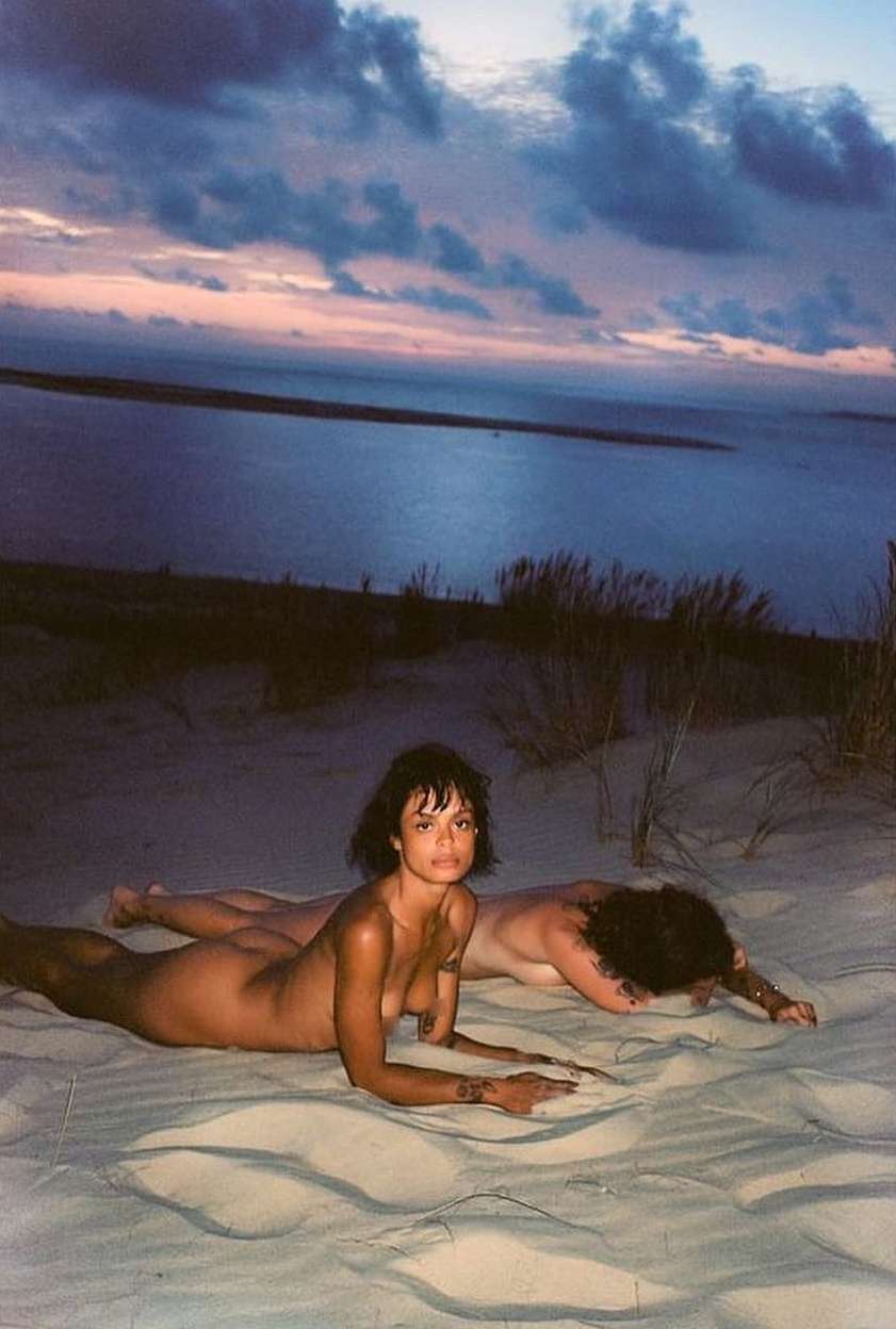 Two people lying prone on a sandy dune overlooking a dim coastline under a twilight sky by Eleonore Wismes