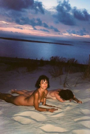 Two people lying prone on a sandy dune overlooking a dim coastline under a twilight sky by Eleonore Wismes