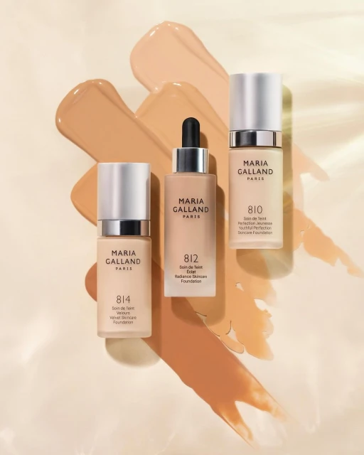 Three bottles of Maria Galland skincare foundation with various shades of the foundation as smears in the background, indoor setting.