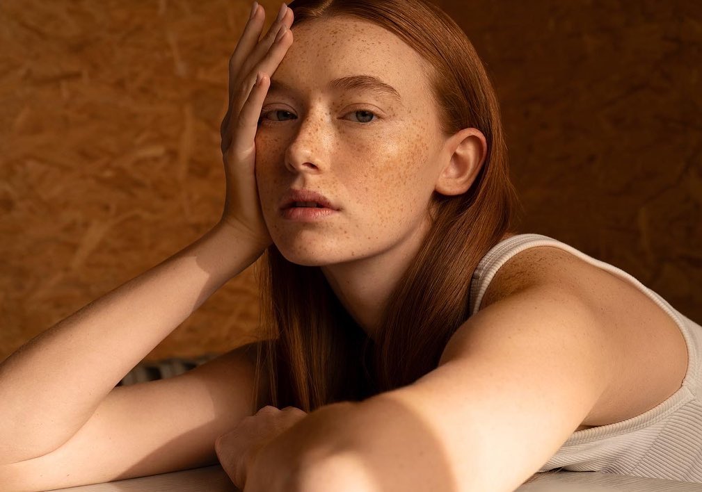 Close-up studio portrait of a red-haired woman with freckles, resting her head on her hand, set against a wood panel background.
