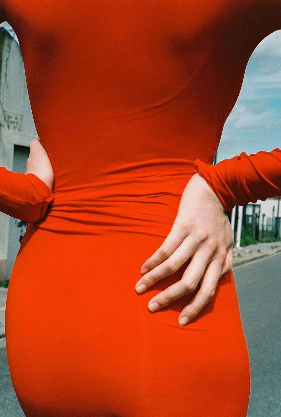 Close-up of a person wearing a fitted long-sleeve red dress with hands on hips, photographed outdoors on an urban street.