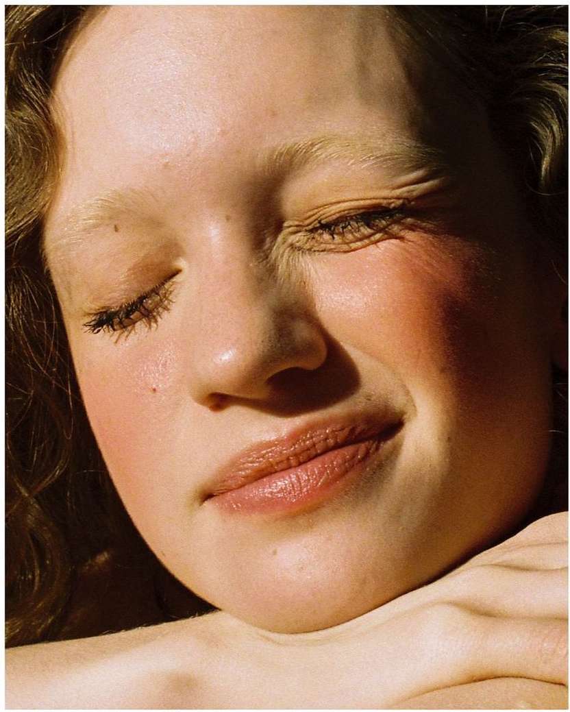 Close-up portrait of a young woman with fair skin, light brown hair, and closed eyes, bathed in soft, warm light highlighting her features.