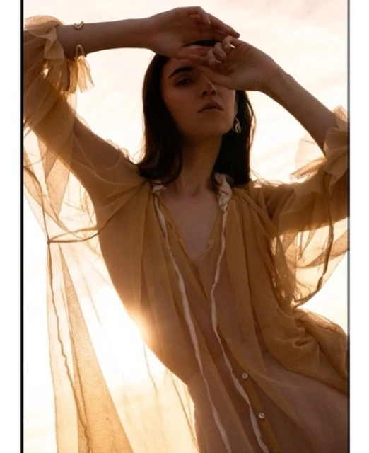 Young woman wearing a flowing sheer gold blouse shields her face from sunlight. She has dark hair, light skin, and a neutral expression.
