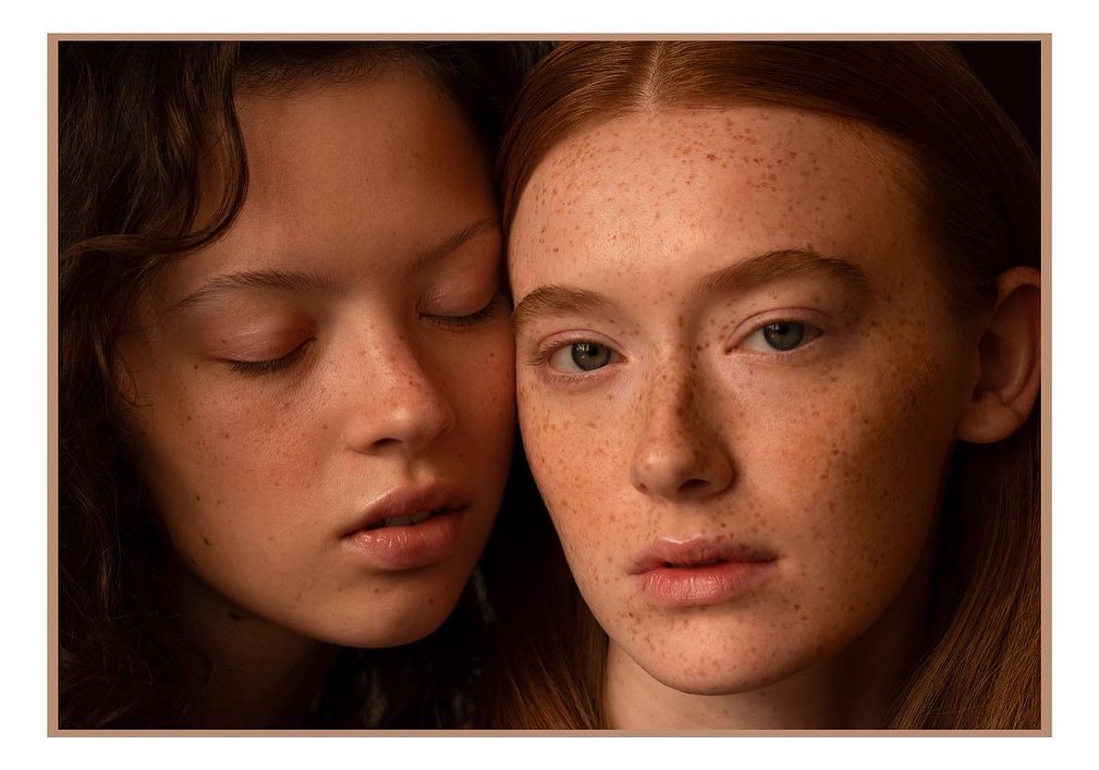Close-up portrait of two young women with freckles, one with red hair and one with dark curly hair, in a softly lit, neutral setting.