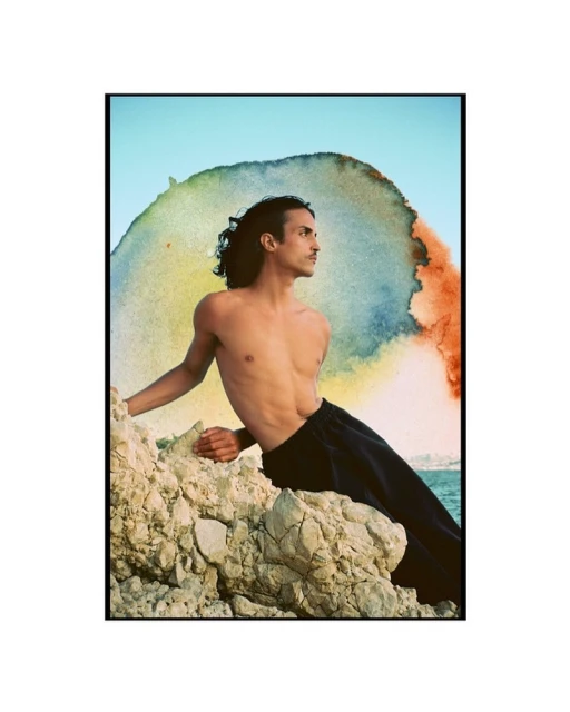 A shirtless adult male with black pants poses on rocks against a watercolor-style background and a bright blue sky in an outdoor setting.