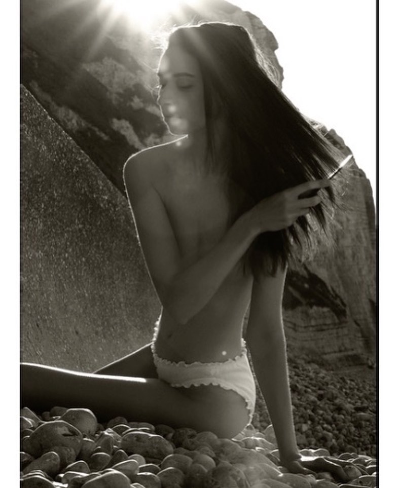Monochrome portrait of a slim woman in underwear seated on a rocky beach, with sunlight filtering through a coastal rock formation.
