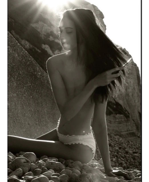 Monochrome portrait of a slim woman in underwear seated on a rocky beach, with sunlight filtering through a coastal rock formation.