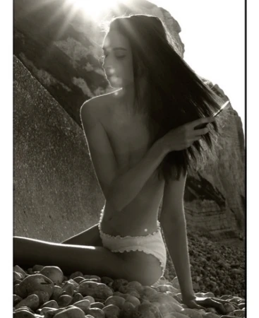 Monochrome portrait of a slim woman in underwear seated on a rocky beach, with sunlight filtering through a coastal rock formation.