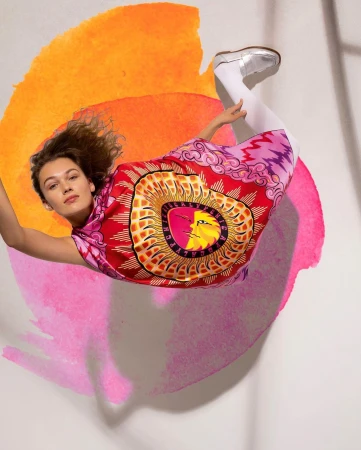 Model wearing a vibrant red graphic dress lying on a painted background with orange and pink watercolor washes.