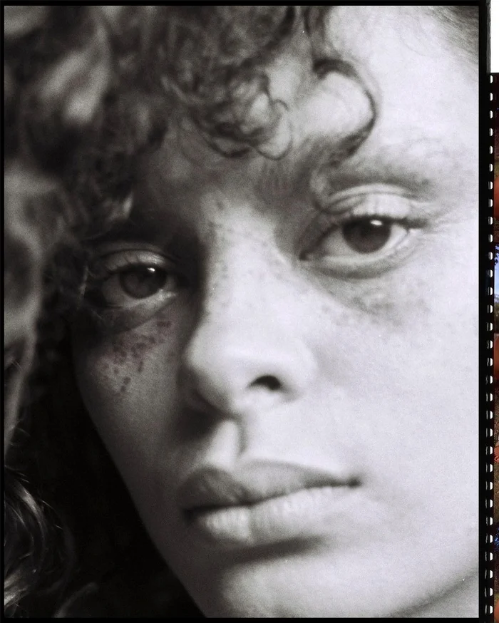 Close-up black and white portrait of a person with curly hair and visible freckles photographed by Eleonore Wismes.