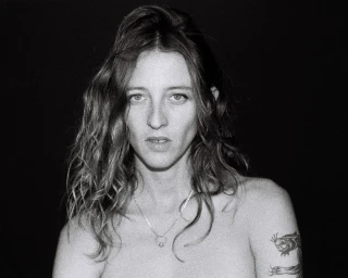 Portrait of a woman with long wavy hair, a silver necklace, and a panther tattoo on her arm against a black background.