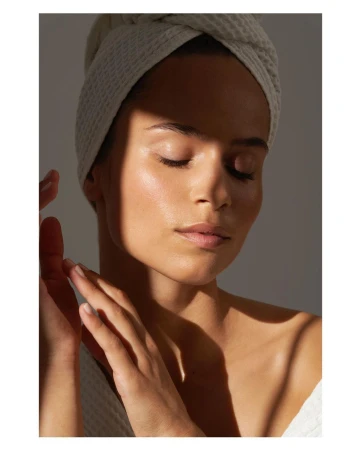 Close-up portrait of a woman with a towel turban and raised hands, illuminated by patterned sunlight against a grey background.