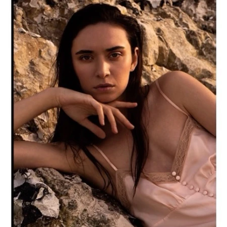 Woman in pink slip dress reclining against rocky coastal background photographed by Eleonore Wismes