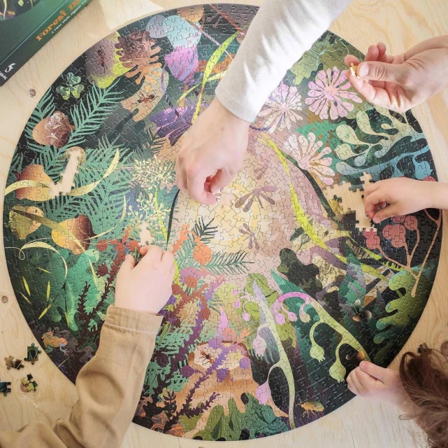 Overhead shot of three people working on a circular jigsaw puzzle with a forest theme on a wooden table.