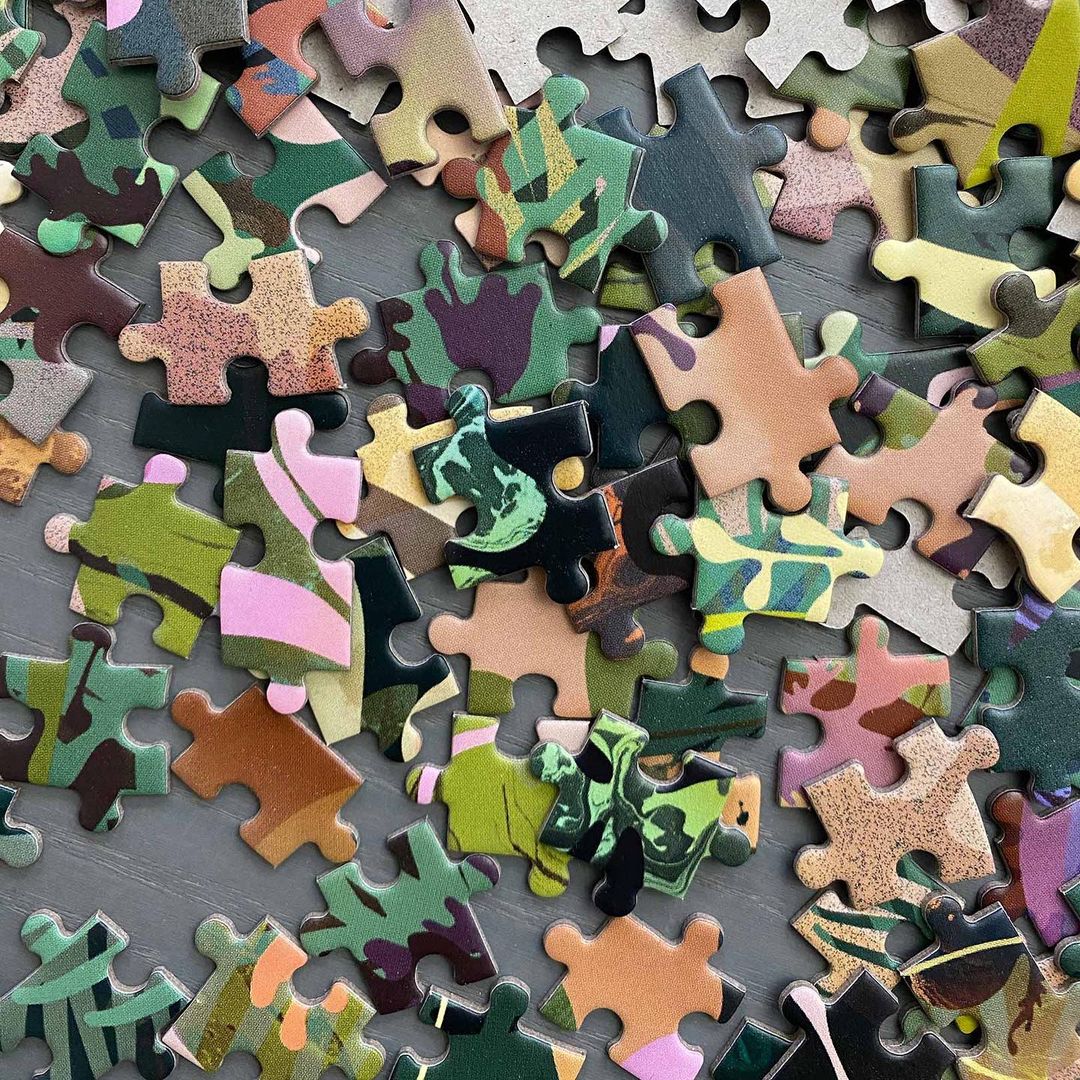 Close-up of a high-angle shot features puzzle pieces with camouflage patterns scattered across a gray surface, creating a textured abstract.
