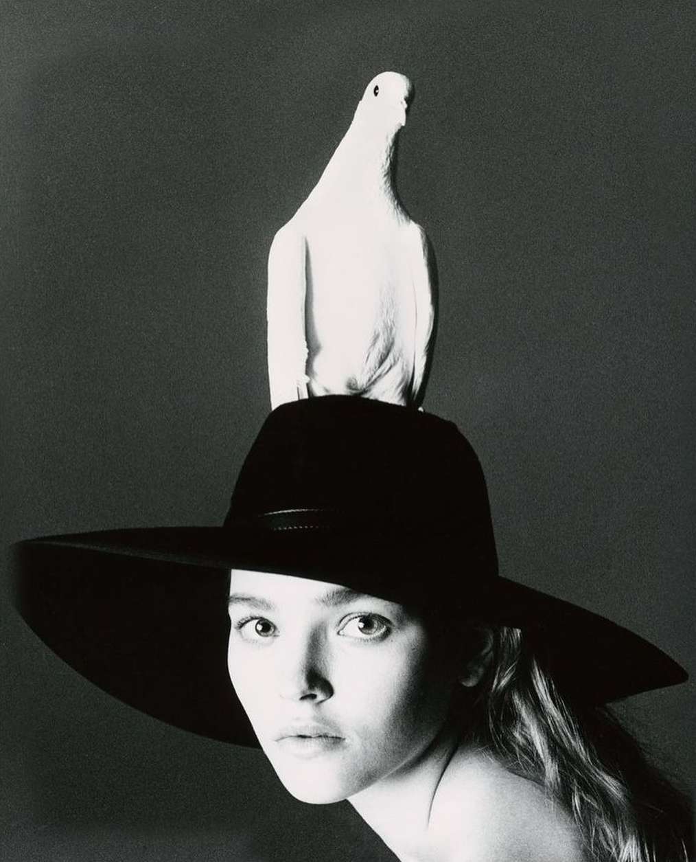Black and white portrait of a woman wearing a wide-brimmed hat with a white dove perched on top, looking directly at the camera.