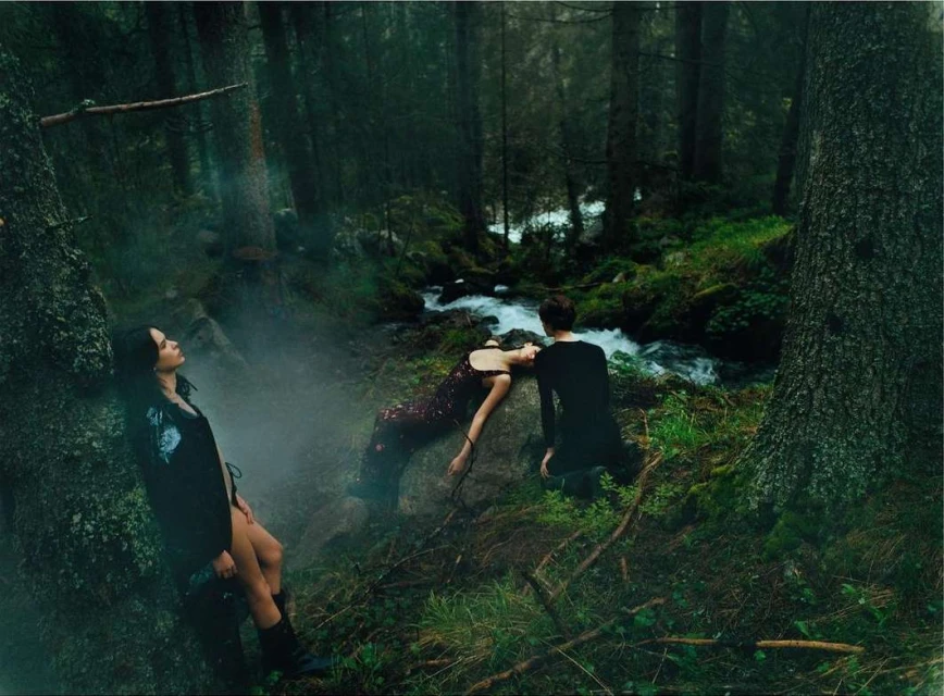 Three people in a forest near a stream. One woman leans against a tree, another lays on a rock, and one person kneels in black clothing.