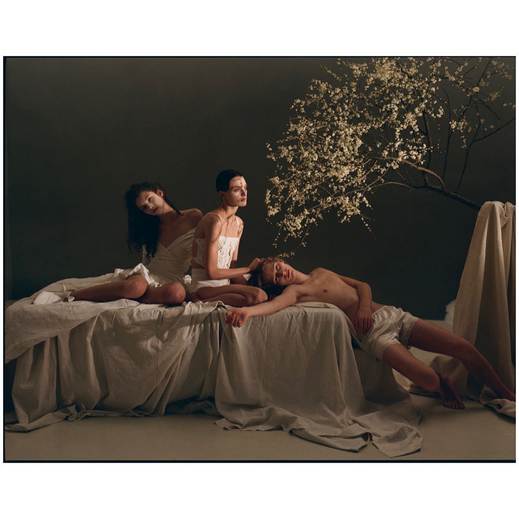 Three figures pose on a bed covered with a white sheet; a blossoming tree branch extends from the right against a grey backdrop.