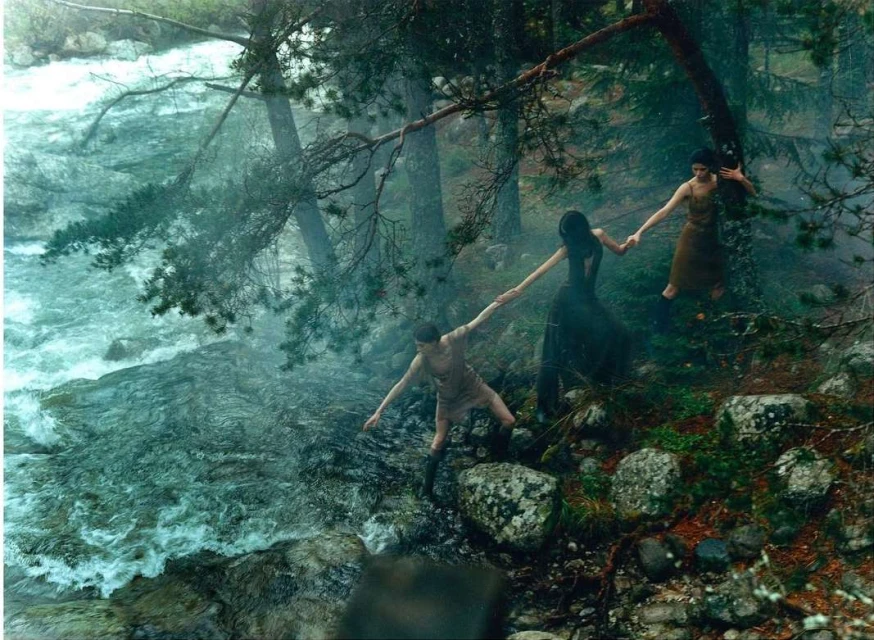 Three figures hold hands near a stream in a forest. One figure stands on the stream's edge, and the others stand further up the bank.