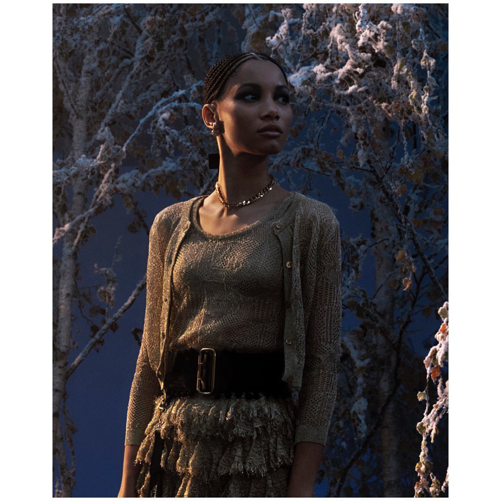Elegant woman wearing a gold knit top, cardigan, and ruffled skirt with a black belt, posing in front of a snow-covered tree.