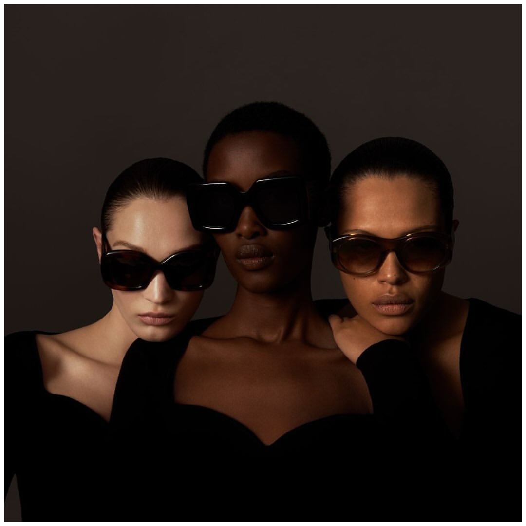 Three women wearing sunglasses in a studio setting. Close-up shot features the diverse trio with dark clothing against a dark background.