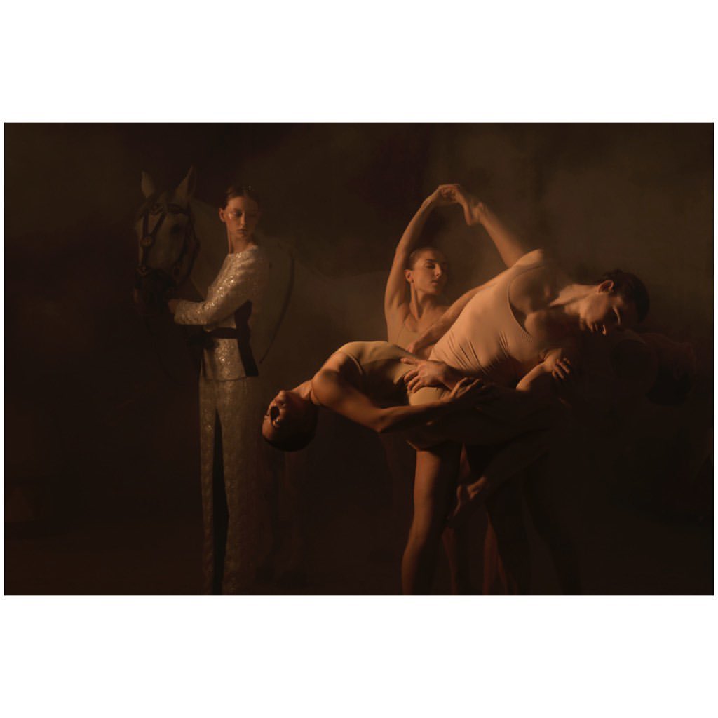 Ethereal image of a woman in a sequined suit holding a horse, juxtaposed with dancers in nude unitards, creating a surreal and artistic composition.