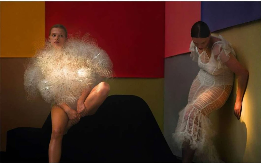 Two fashion models pose indoors against colorful wall panels, wearing unique textured couture dresses, one with a dandelion-like texture.