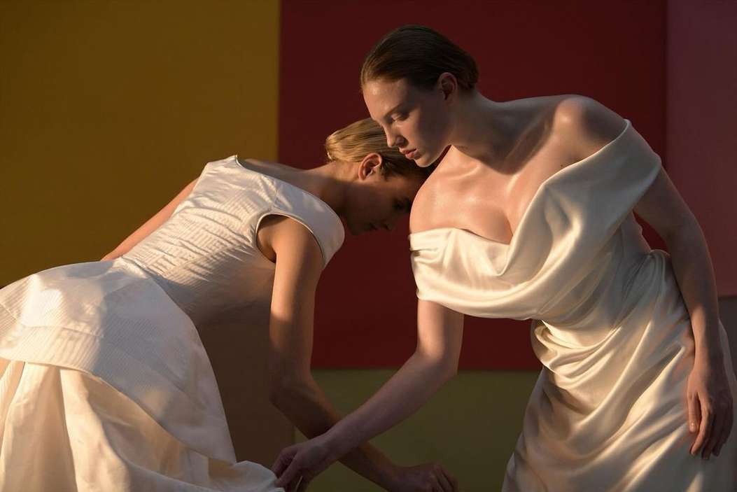 Two adult females in flowing white dresses pose together in a studio with a vibrant color-blocked background featuring yellow, red, and pink.