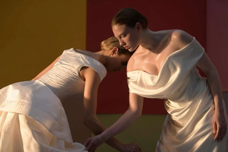 Two adult females in flowing white dresses pose together in a studio with a vibrant color-blocked background featuring yellow, red, and pink.