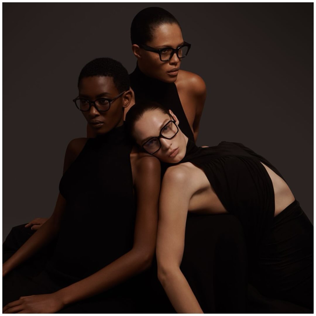 Three models wearing black sleeveless tops and black eyeglasses posing together in a studio setting.