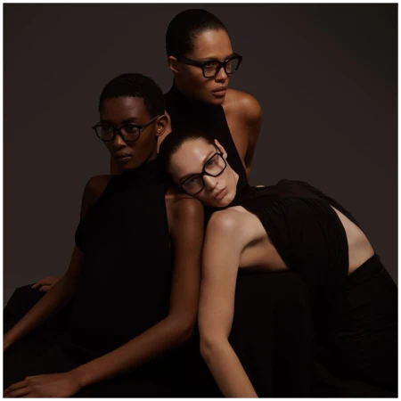 Three models wearing black sleeveless tops and black eyeglasses posing together in a studio setting.