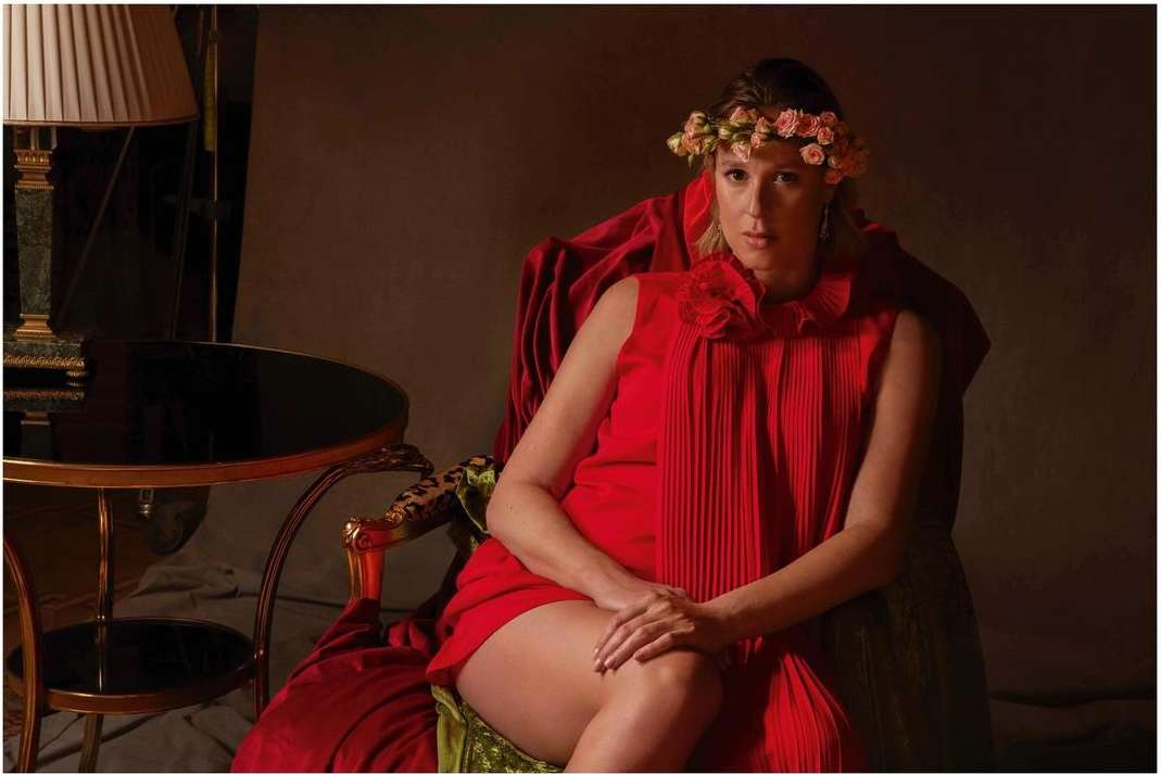 Adult woman in red dress sits on a velvet-covered chair, accented with gold trim, and wears a floral headpiece in a dimly lit room.