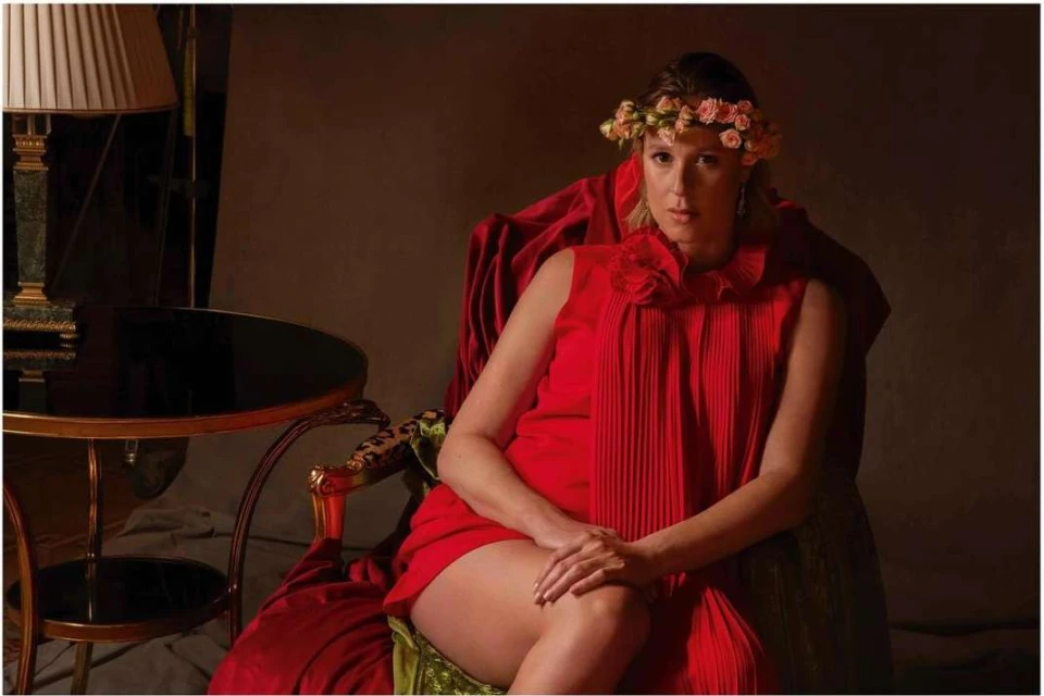 Adult woman in red dress sits on a velvet-covered chair, accented with gold trim, and wears a floral headpiece in a dimly lit room.