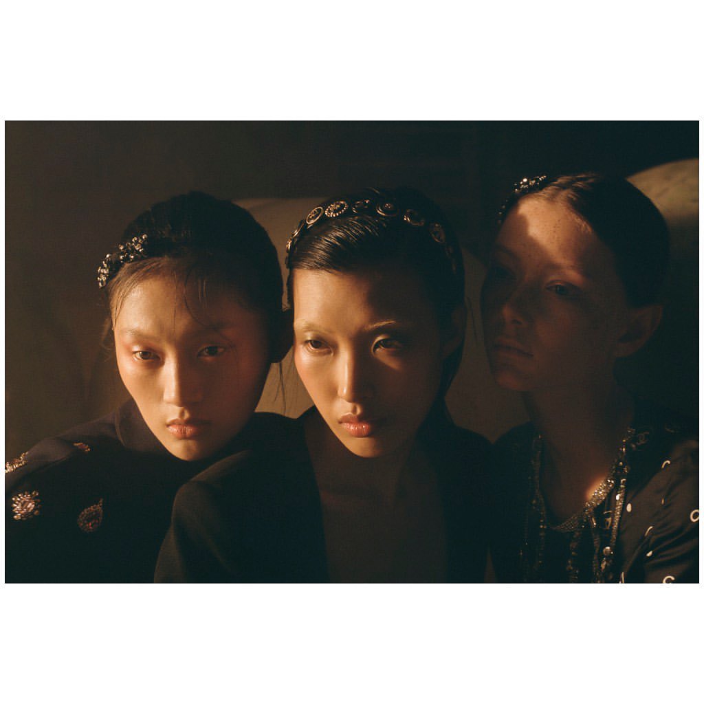 Three young adult women pose together wearing headbands and black jackets in a studio with dramatic lighting.