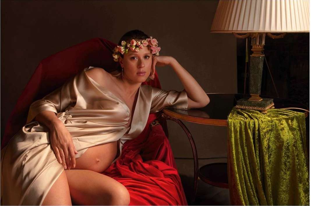 Pregnant woman in a gold wrap poses in a red velvet chair with a floral crown and lamp with a green velvet drape.