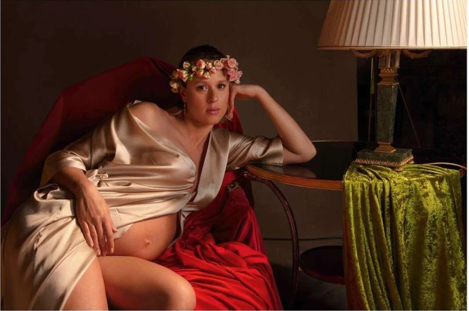 Pregnant woman in a gold wrap poses in a red velvet chair with a floral crown and lamp with a green velvet drape.