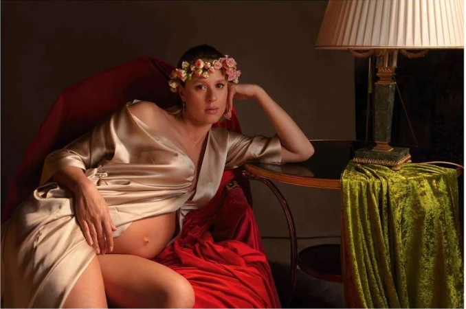 Pregnant woman in a gold wrap poses in a red velvet chair with a floral crown and lamp with a green velvet drape.