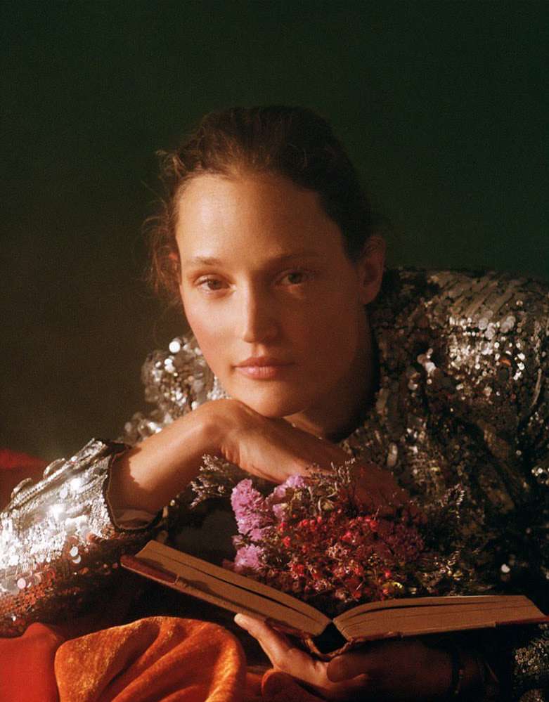 Portrait of a young woman with flowers in a book, wearing a sparkly top, in front of a dark green background, eye-level shot.