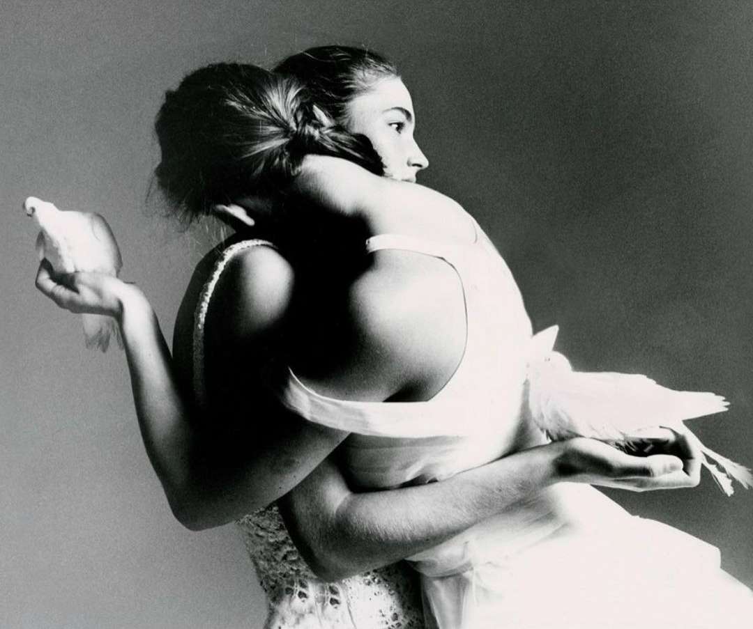 Monochrome portrait of two women in white dresses embracing while holding white doves, soft lighting, and a simple dark background.
