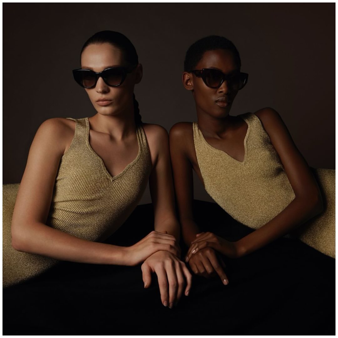 Two fashion models pose indoors wearing gold tank tops and sunglasses in a medium shot with low-key lighting and a neutral background.