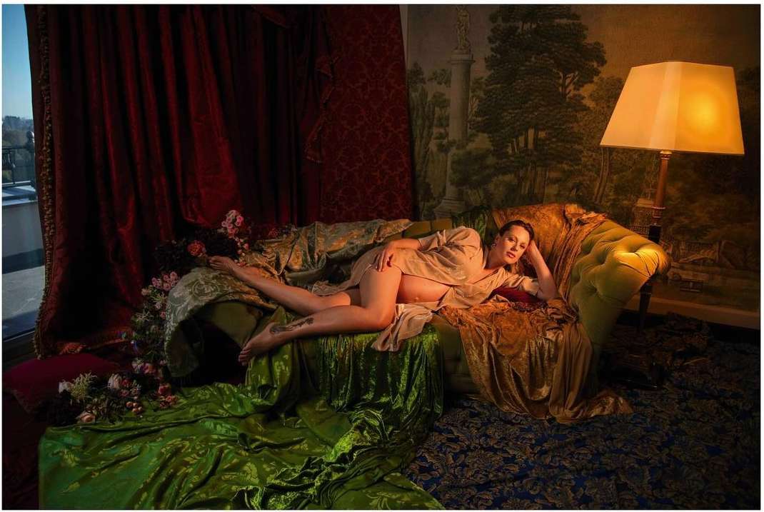 Woman reclines on a green velvet couch in front of red drapes and a wall with mural art illuminated by a floor lamp.