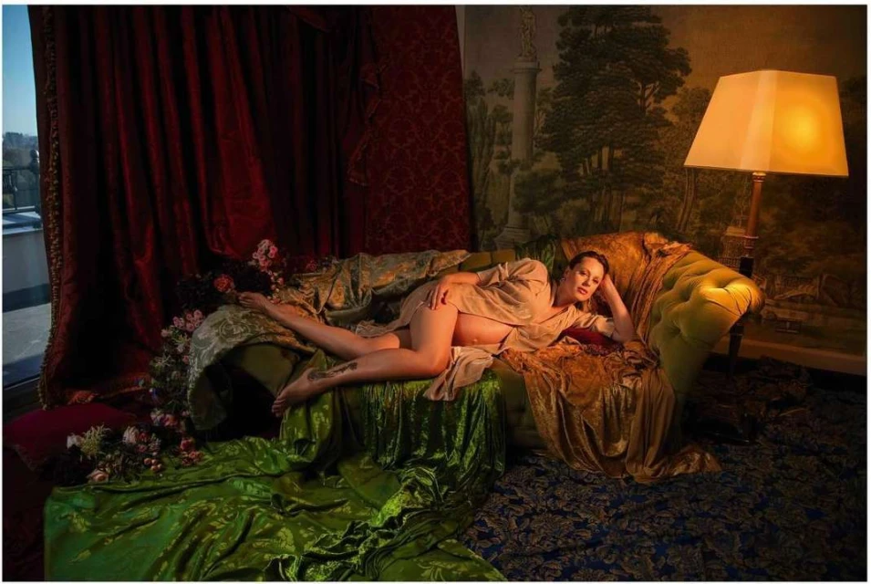 Woman reclines on a green velvet couch in front of red drapes and a wall with mural art illuminated by a floor lamp.